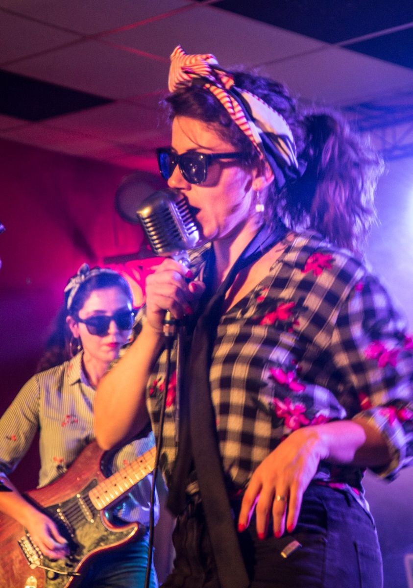 Lasimi canta in un microfono vintage, con bandana e occhiali da sole, una chitarrista alle spalle