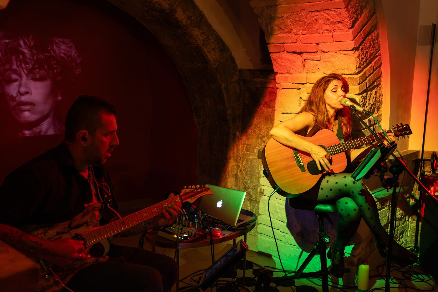 Lasimi canta alla chitarra acustica accanto ad Andrea Frittelli, con un volto proiettato sul muro